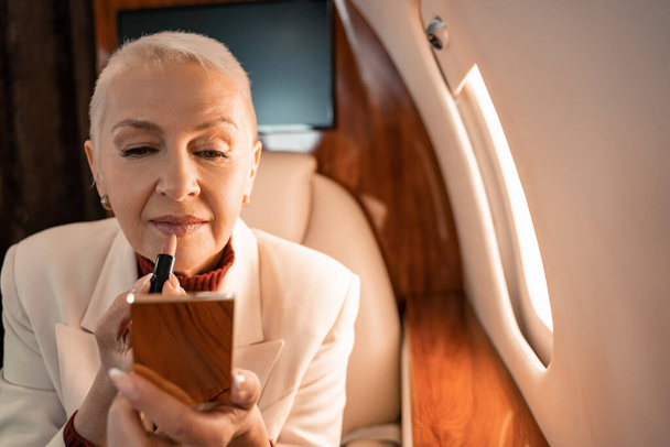 Femme mature tenant rouge à lèvres et miroir flou dans un avion privé  - Photo, image
