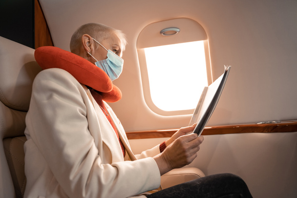Geschäftsfrau mit medizinischer Maske und Nackenkissen liest Magazin im Flugzeug  - Foto, Bild