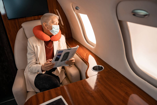 Geschäftsfrau mit Nackenkissen und medizinischer Maske hält Magazin in der Hand und blickt auf Flugzeugfenster  - Foto, Bild