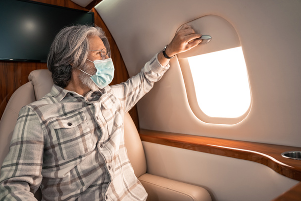 Άντρας με προστατευτική μάσκα κοιτάζει το παράθυρο σε ιδιωτικό αεροπλάνο  - Φωτογραφία, εικόνα