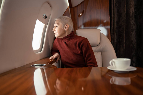 Mature woman looking at window of private jet near blurred cup of coffee  - Foto, Bild