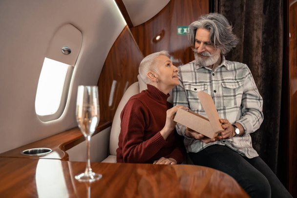 Usmívající se žena drží dárek v blízkosti manžela a rozmazané šampaňské v letadle  - Fotografie, Obrázek