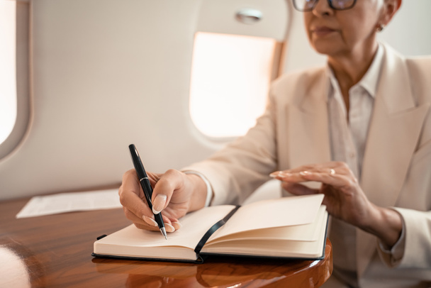 Vista recortada de la mujer de negocios borrosa escribiendo en el cuaderno en jet privado  - Foto, Imagen