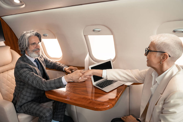 Mosolygó üzletember kezet rázott üzletasszonnyal laptopok közelében a repülőn  - Fotó, kép