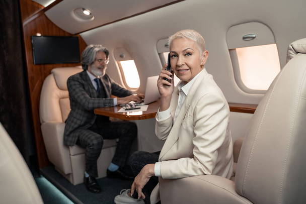 Mujer de negocios sonriente hablando en el teléfono inteligente y mirando a la cámara cerca borrosa empresario en avión  - Foto, Imagen