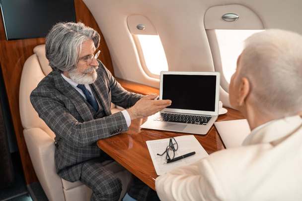 Geschäftsmann zeigt auf Laptop neben Papier und verschwommene Geschäftsfrau im Privatflugzeug  - Foto, Bild