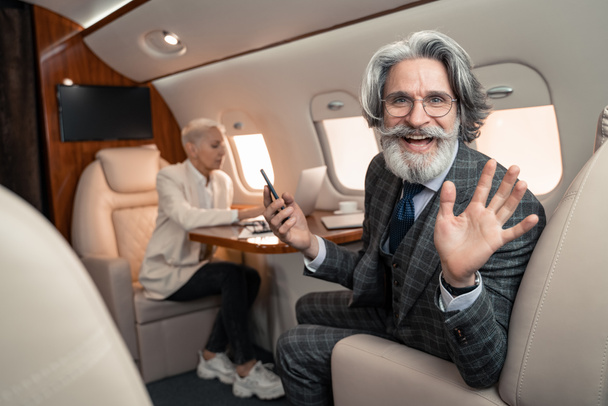 Hombre de negocios sonriente con teléfono inteligente saludando a la cámara cerca de la mujer de negocios borrosa en avión  - Foto, Imagen