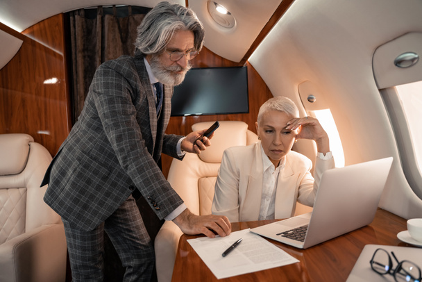 Geschäftsmann mit Smartphone schaut im Privatflugzeug auf Papier neben Geschäftsfrau mit Laptop  - Foto, Bild