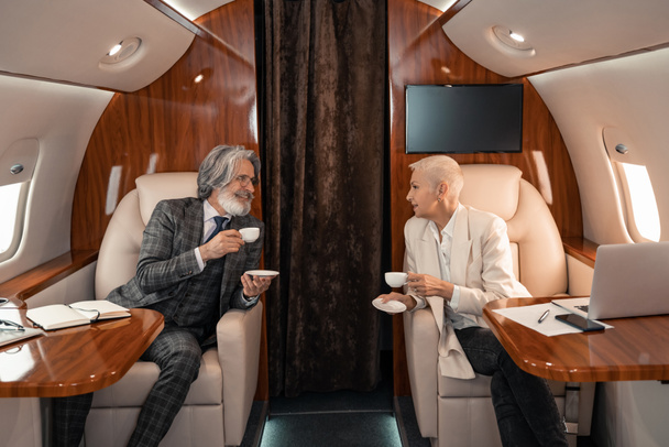 Homme d'affaires souriant tenant un café près d'une femme d'affaires et des gadgets dans l'avion  - Photo, image