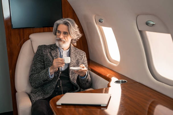 Uomo d'affari in possesso di caffè vicino computer portatile offuscata in aereo privato  - Foto, immagini
