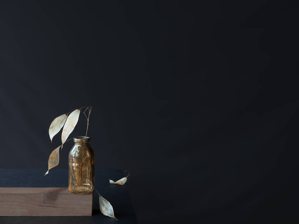 Trendy monochrome stilleven zilver gedroogde tak van lunaria in transparante gouden lege flesje, ruw geschilderd, gestripte verf, 2 bladeren op tafel. Op zwarte achtergrond, horizontale banner, kopieer ruimte - Foto, afbeelding