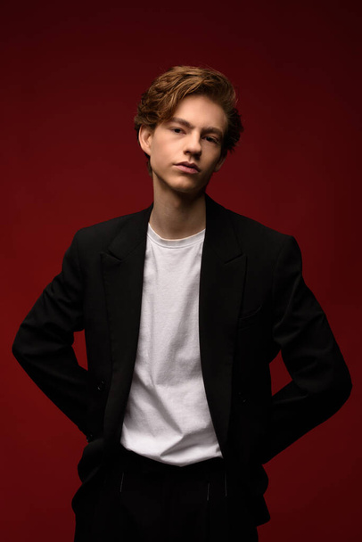 Stylish young guy with gorgeous blond hair and beautiful skin in a black jacket and white T-shirt. guy smiling and posing with his hands on a red background - Foto, afbeelding