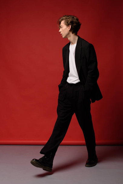 Stylish young guy with lush blond hair and beautiful skin in a black jacket, pants and white T-shirt. He are goes beautifully wide step in bl ack shoes on a red background. hands in pockets - Фото, изображение