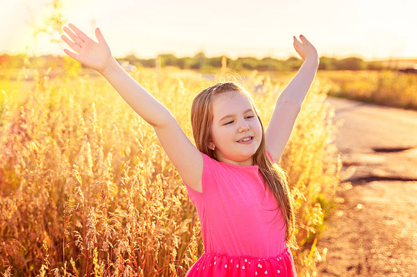 Une petite fille souriante aux cheveux longs dans une robe rose reste le long d'une route de campagne. Bonne enfance, mode de vie, humeur chaleureuse, vacances d'été. concept - Photo, image