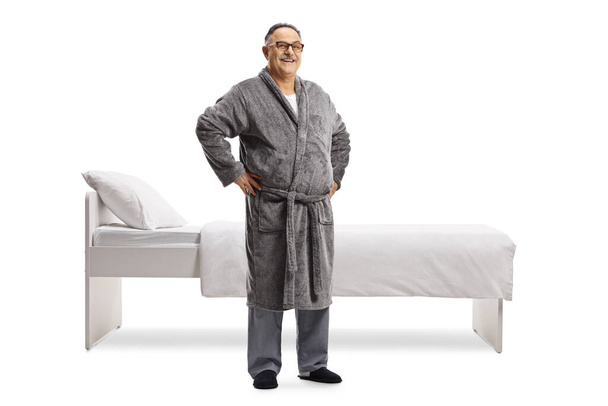 Full length portrait of a mature man in pajamas and a robe standing in front of a bed isolated on white background - Fotoğraf, Görsel