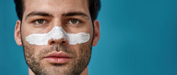 Cropped portrait of serious brunette guy with white mask applied on nose for cleansing pores looking away, posing isolated over blue background - Foto, immagini