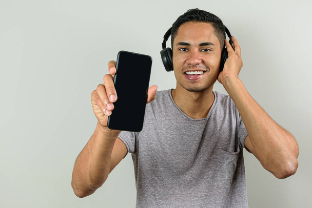 Hymyilevä 28-vuotias brasilialainen yhdellä kädellä langattomassa kuulokkeessa, ja toinen osoittaa älypuhelimensa kameralle. - Valokuva, kuva