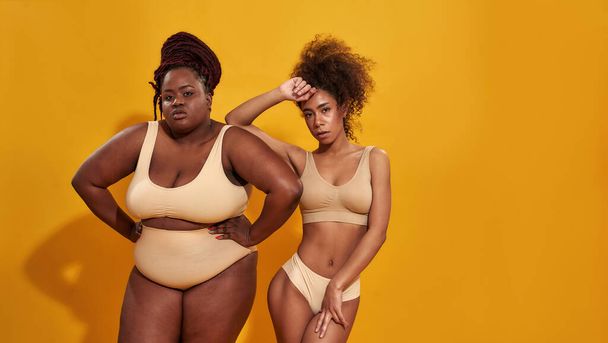 Studio shot of two confident plus size and slim african american women in beige underwear looking at camera while posing together isolated over yellow background - Foto, Imagen