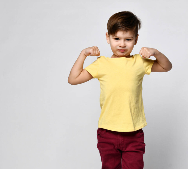 Handsome confident kid wearing casual yellow t-shirt smiles happily and raises clenched fists, tightening muscles, feeling strong and full of energy. - Фото, изображение
