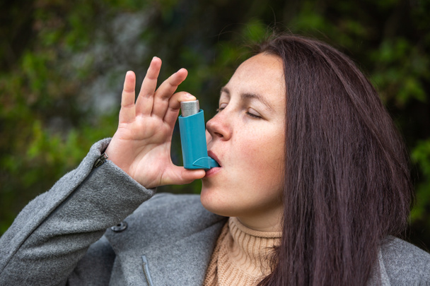 Bella giovane donna bruna che utilizza un inalatore di asma durante un forte attacco di asma, il prodotto farmaceutico viene utilizzato per prevenire e trattare respiro affannoso e mancanza di respiro, concetto di assistenza sanitaria - Foto, immagini