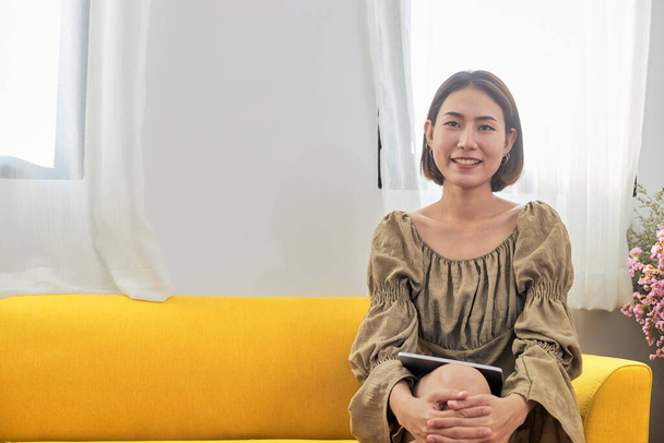 Портрет счастливой азиатки, сидящей на жёлтом диване возле окна на случайном отдыхе дома. - Фото, изображение