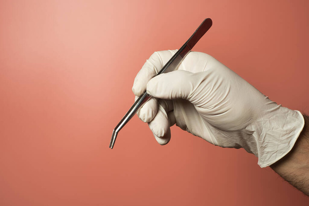 Unrecognized man wearing latex gloves holding dental tweezers against a light red background. - Фото, зображення