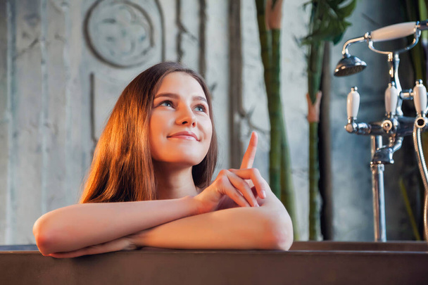 Portrait of young cute cheerful woman in bathtub shows finge and looking away over old interior bathroom. Advertising concept of healthy lifestyle and self care. Copy space for site or SPA - Foto, Bild