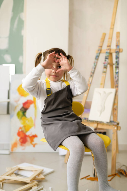 lapsi, pieni tyttö, jolla on poninhäntä, nuori taiteilija kotona studio-työpajassa, joka näyttää käsillään rakkauden-sydämen symbolin, keskittyy käsiinsä - Valokuva, kuva