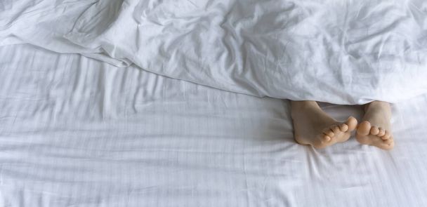 朝起きてる。朝、ベッドのカバーの下の足。白いベッドの下の女性の足。リラックス、睡眠、休息の概念. - 写真・画像
