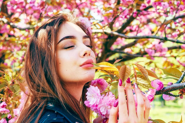 Young beautiful girl with long hair enjoys the beauty of spring nature near blossoming sakura tree - Фото, изображение