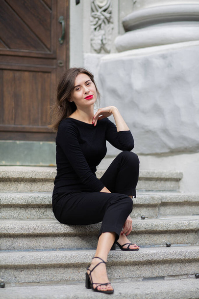 Lifestyle shot d'une belle jeune femme aux lèvres rouges et aux cheveux bouclés courts porte une tenue noire tendance assise dans les escaliers - Photo, image