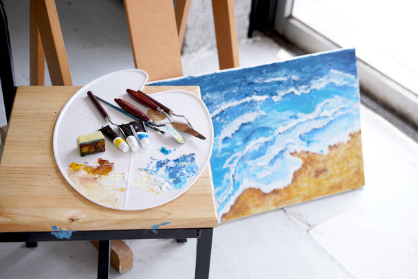 Akrylové moře a plážové malby na kreslicím stojanu s paletou a štětcem. Modrá vlna na zlaté pískové ruční malbě štětcem a paletovou technikou nože. Umění na plátně. - Fotografie, Obrázek