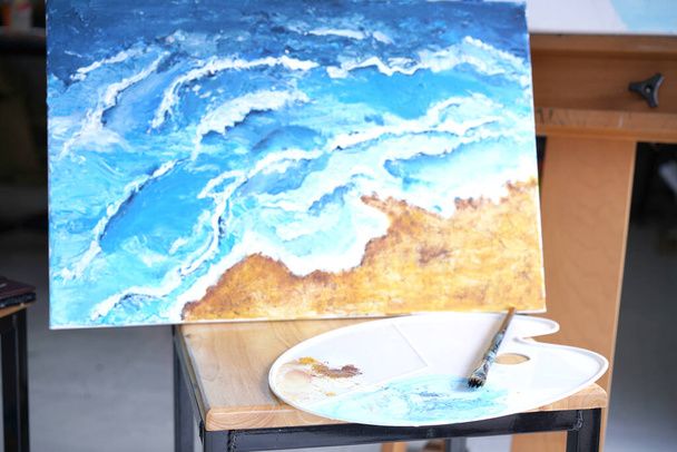 Акриловое море и пляжная живопись на стенде с палитрой и кистью. Голубая волна на золотистом песке, роспись кистью и ножом в технике палитры. Картины на холсте. - Фото, изображение