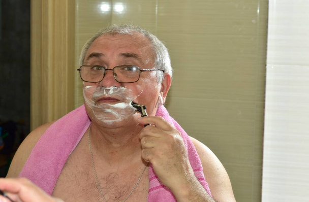 Ο άντρας ξυρίζει το αριστερό του μάγουλο κοντά στα χείλη του ενώ κοιτά στον καθρέφτη.. - Φωτογραφία, εικόνα