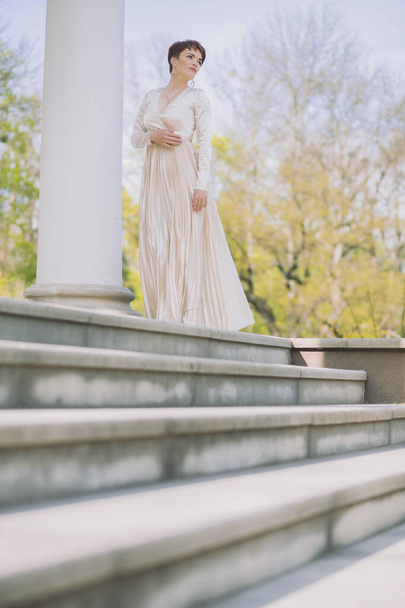 όμορφη λεπτή γυναίκα σε ένα μακρύ βραδινό φόρεμα στέκεται στις στήλες - Φωτογραφία, εικόνα