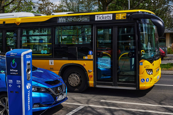 Berlim, Alemanha - 8 de maio de 2021: Um carro elétrico ecológico em uma estação de carregamento e um ônibus de transporte público que estão um pouco fora de foco. O foco está no fundo através das janelas do ônibus. - Foto, Imagem