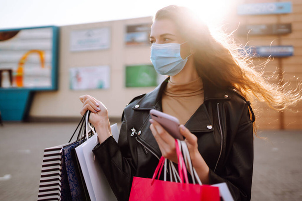 Smiling woman in protective medical mask with shopping bags using her phone. Young woman after shopping on the city street. Online shopping concept. Shopping in the coronavirus epidemic.  - Photo, image
