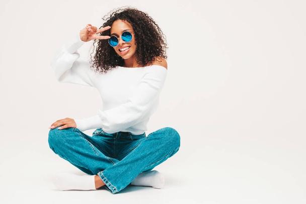 Beautiful black woman with afro curls hairstyle.Smiling model in sweater and trendy jeans clothes. Sexy carefree female posing on white background in studio. Tanned and cheerful in sunglasses - Fotografie, Obrázek