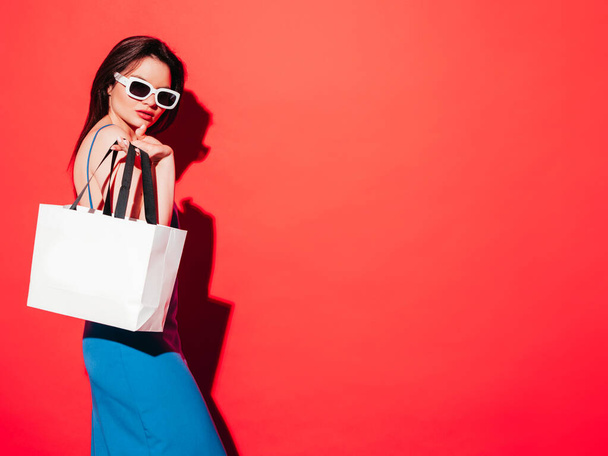 High fashion portrait of young beautiful brunette woman wearing nice blue summer dress.Sexy trendy model posing near red wall in studio.Fashionable female in sunglasses. Holding shopping bag - Photo, image