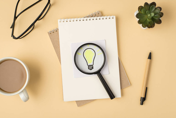 Top view photo of cup glasses flowerpot pen two notebooks and magnifier glass on sticker with light bulb drawing on isolated beige background - Foto, afbeelding