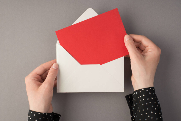 Première personne vue du dessus photo de mains féminines tenant enveloppe blanche ouverte avec carton rouge sur fond gris isolé avec copyspace - Photo, image