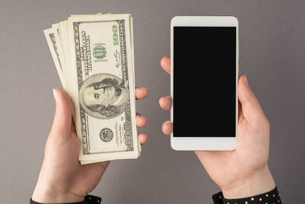 スマートフォンを手にした女性の手の写真と、コピースペース付きの隔離されたグレーの背景にある100ドル紙幣の山 - 写真・画像