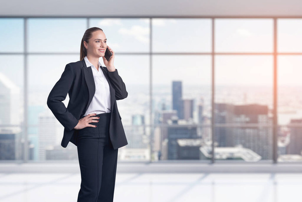 Smiling attractive businesswoman wearing white shirt and black suit is having conference call via smartphone. Panoramic office on background with financial downtown city view. - 写真・画像