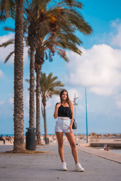 Summer lifestyle, Caucasian brunette with white shorts and black t-shirt on the Mediterranean coast walking next to some palm trees - Foto, afbeelding
