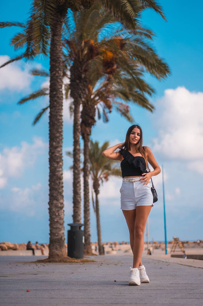 Summer lifestyle, portrait of a young brunette Caucasian woman with white shorts and black t-shirt on the Mediterranean coast next to some palm trees - 写真・画像