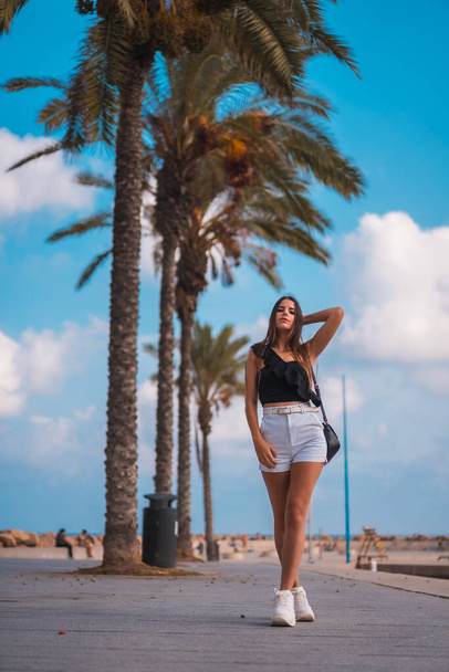 Summer lifestyle, portrait of a young brunette Caucasian woman with white shorts and black t-shirt on the Mediterranean coast next to some palm trees - Фото, изображение