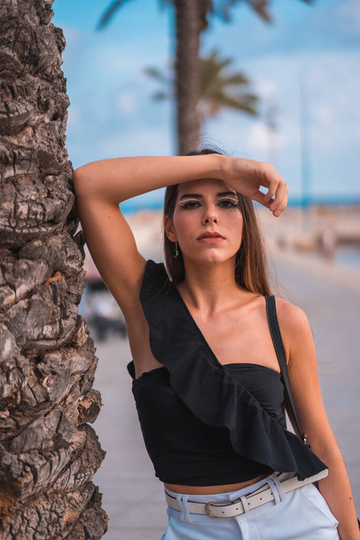 Summer lifestyle, portrait of a young brunette Caucasian woman with white shorts and black t-shirt on the Mediterranean coast next to some palm trees - Foto, Imagem