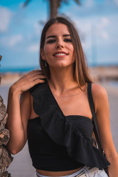 Smile of a young brunette Caucasian with white shorts and black t-shirt on the Mediterranean coast next to some palm trees - 写真・画像