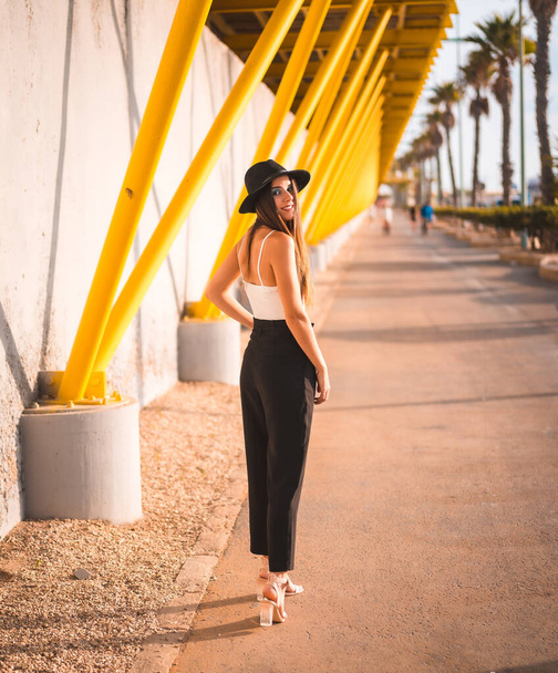 A young brunette in heels, pants and black hat on a yellow boardwalk - Foto, Imagem