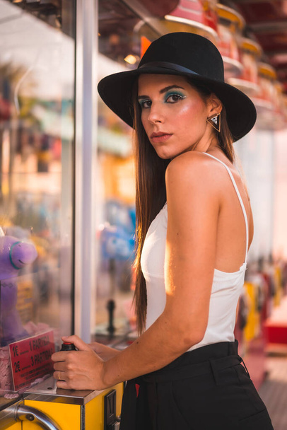 Smile of a young brunette with a black hat and white t-shirt enjoying in an amusement park on an arcade machine in summer - Foto, afbeelding